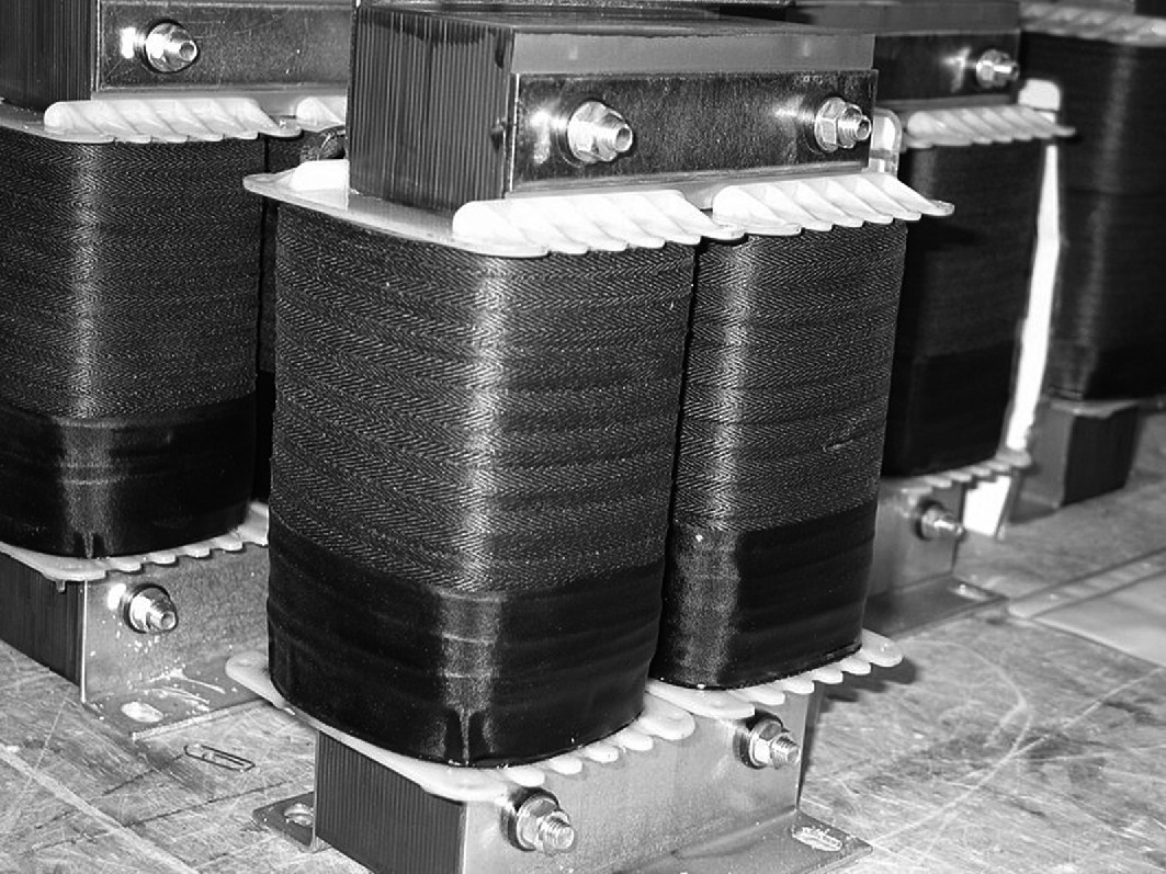 A black and white photo of industrial transformers. The image shows multiple large, rectangular coils with metal components and bolts, arranged in a row. The coils are wrapped in a dark material and mounted on a base, suggesting a setting in a manufacturing or electrical facility.
