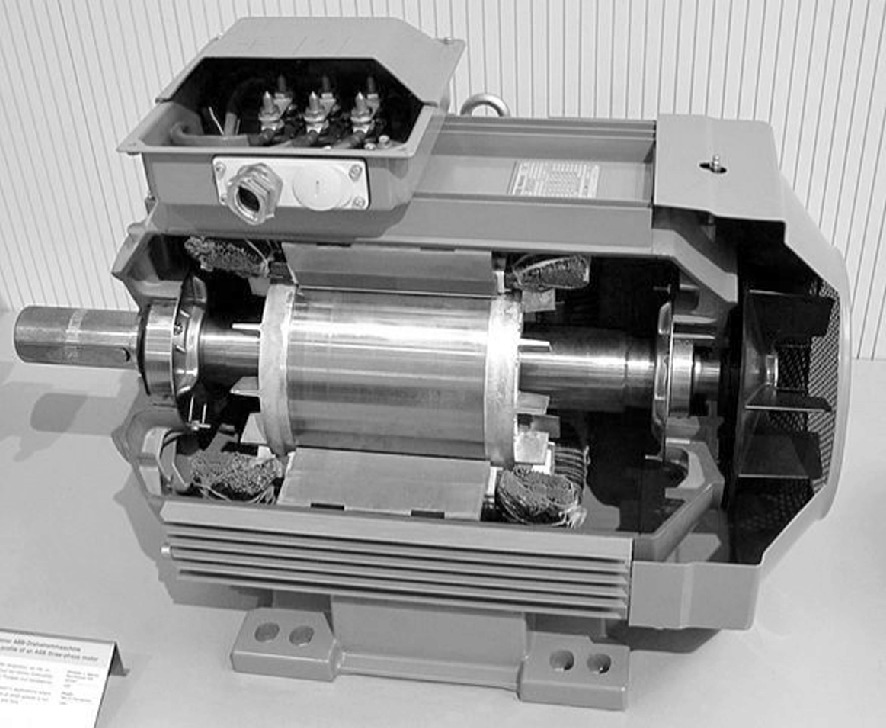A black and white photo of an industrial electric motor with its casing partially removed to reveal the internal components. The motor features a cylindrical rotor and visible wiring connections at the top. The background is a textured surface, and a small label or card is placed near the motor, though the text is not legible.