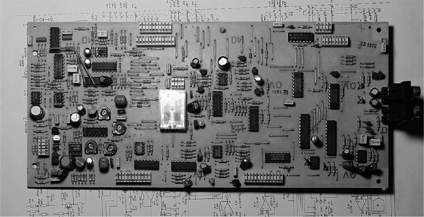 A rectangular circuit board with various electronic components, including resistors, capacitors, integrated circuits, and connectors, is placed on top of a detailed circuit diagram. The diagram underneath shows lines and symbols representing electrical connections and components, providing a schematic view of the circuit board's design.