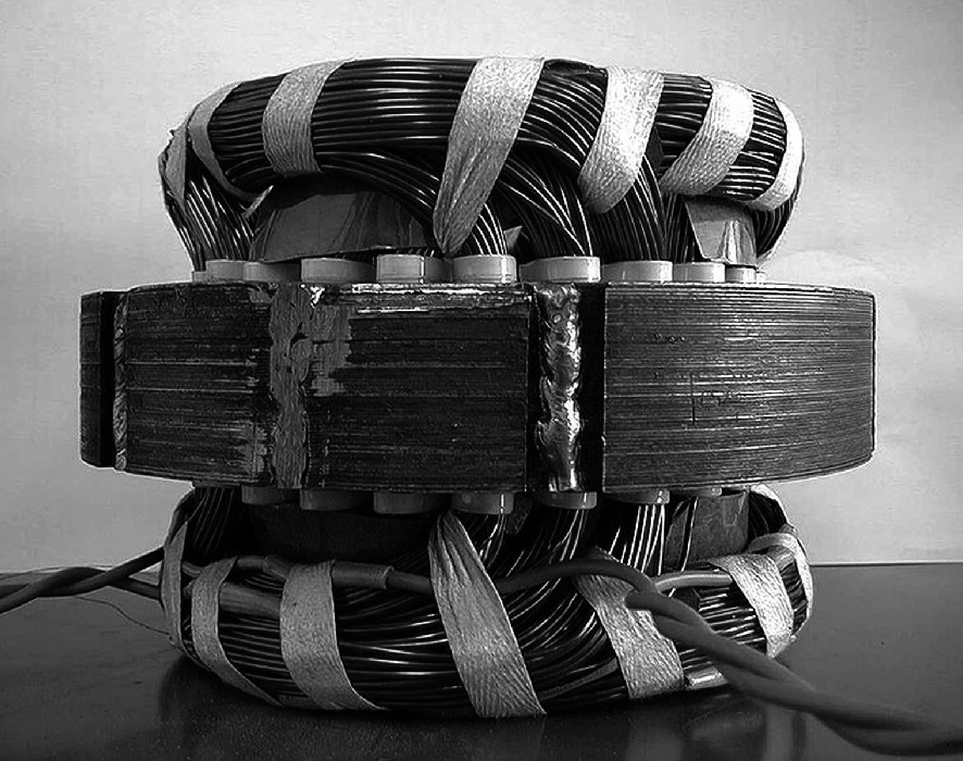 A black and white photo of a large, coiled electrical component, possibly a transformer or inductor. The component is wrapped with thick, striped wires and secured with metal bands. The wires are neatly arranged, and the structure appears robust and industrial. The background is plain, emphasizing the intricate design of the component.