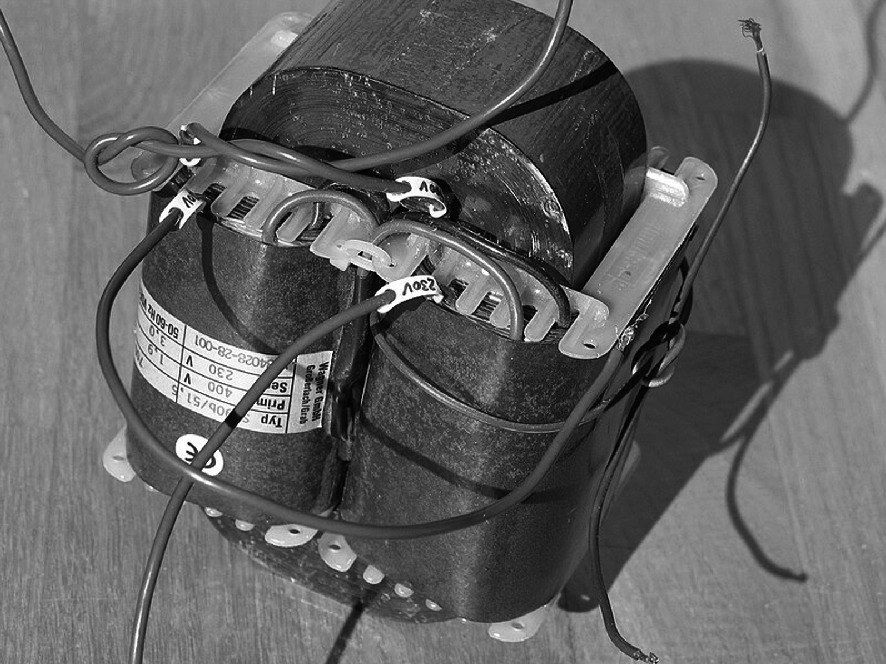 A black and white photo of an electrical transformer with visible coils and wires extending from the top. The transformer is mounted on a base, and there is a label with text on one side, though the text is not legible. The image is taken from an overhead angle, showing the structure and connections of the device.