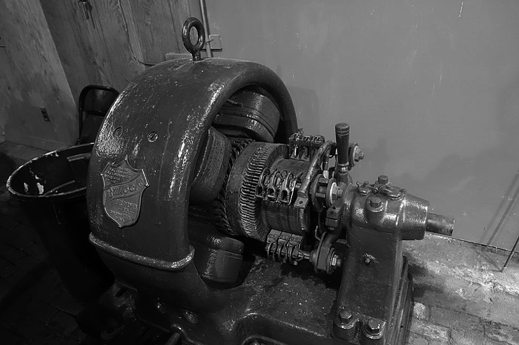A vintage industrial machine with visible gears and a metal casing, positioned in a workshop setting. The machine appears to be used for mechanical processes, with intricate components and a sturdy build. The background includes a plain wall, adding to the industrial atmosphere.